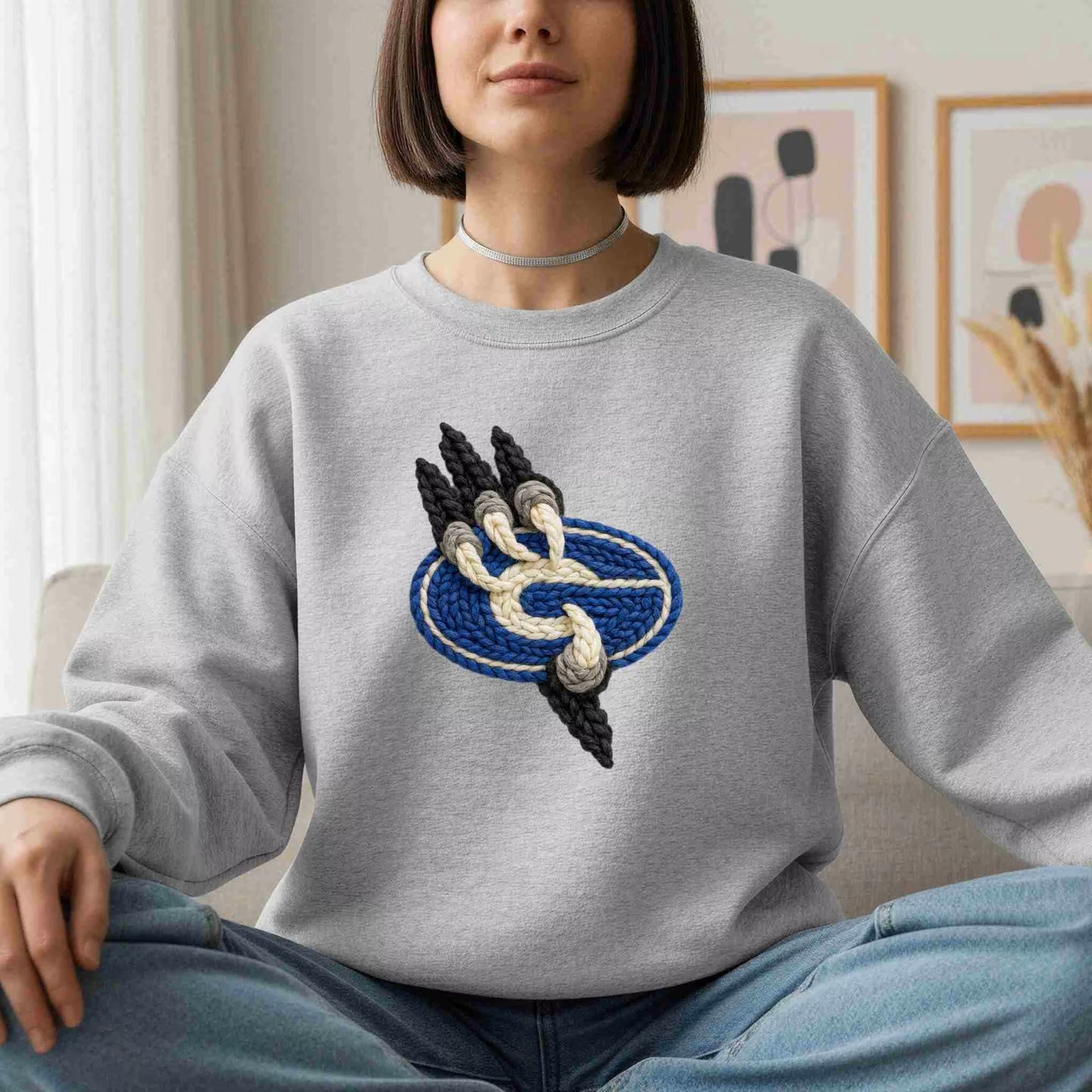 A woman sits cross-legged indoors wearing the Gateway Griffins Crewneck by Becca's Banging Designs, featuring a blue oval and knot design. With chin-length hair, a simple choker, and abstract art behind her, she exudes cozy style.