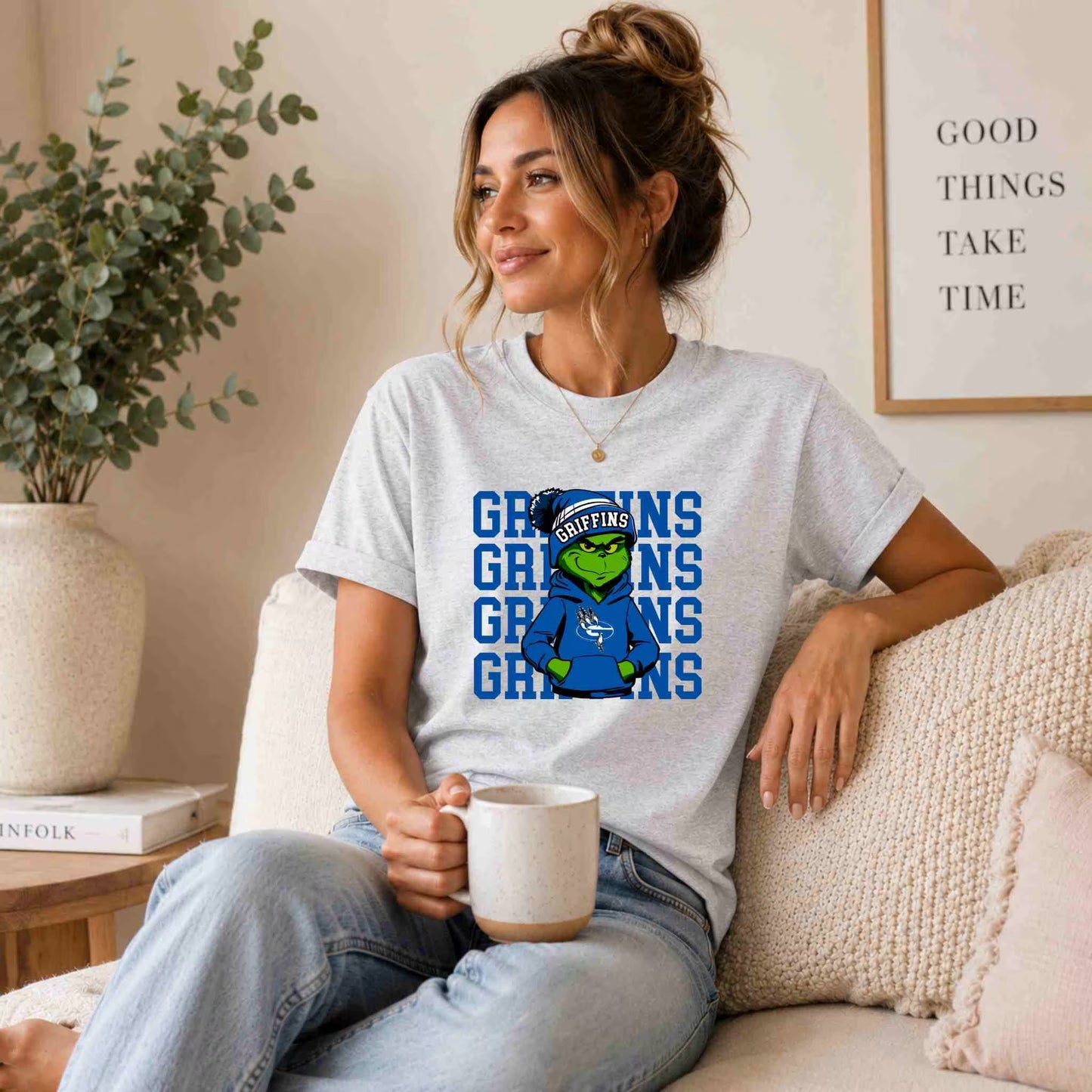 A woman with a casual hairstyle sits on a beige couch, smiling and holding a mug. She wears the Becca's Banging Designs Griffins Graphic Tee Youth Comfort and School Spirit with a cartoon mascot and blue text. A plant and framed quote are behind her.