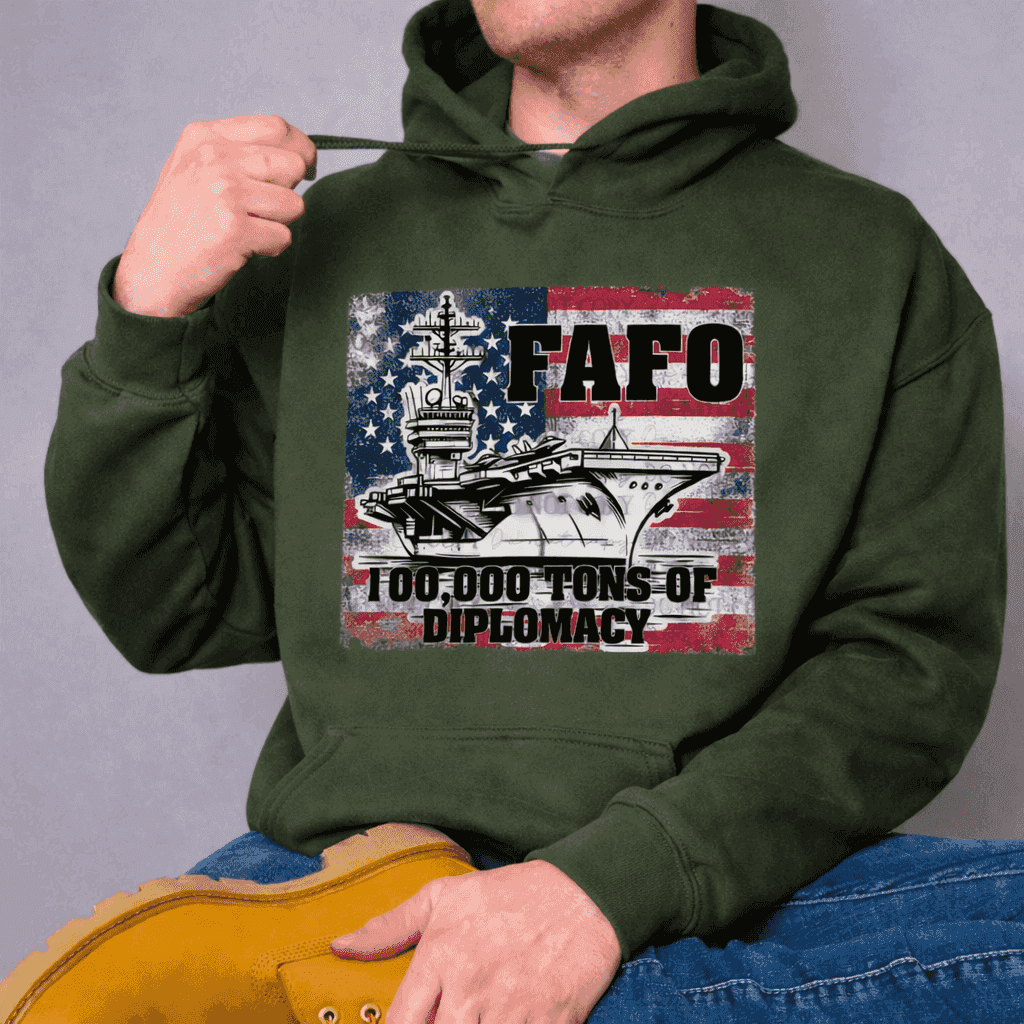 Wearing the FAFO Diplomacy | Hoodie by Becca's Banging Designs LLC, featuring a green color, aircraft carrier print, distressed U.S. flag, and "100,000 Tons of Diplomacy," a person sits cross-legged and tugs the hood’s drawstring.