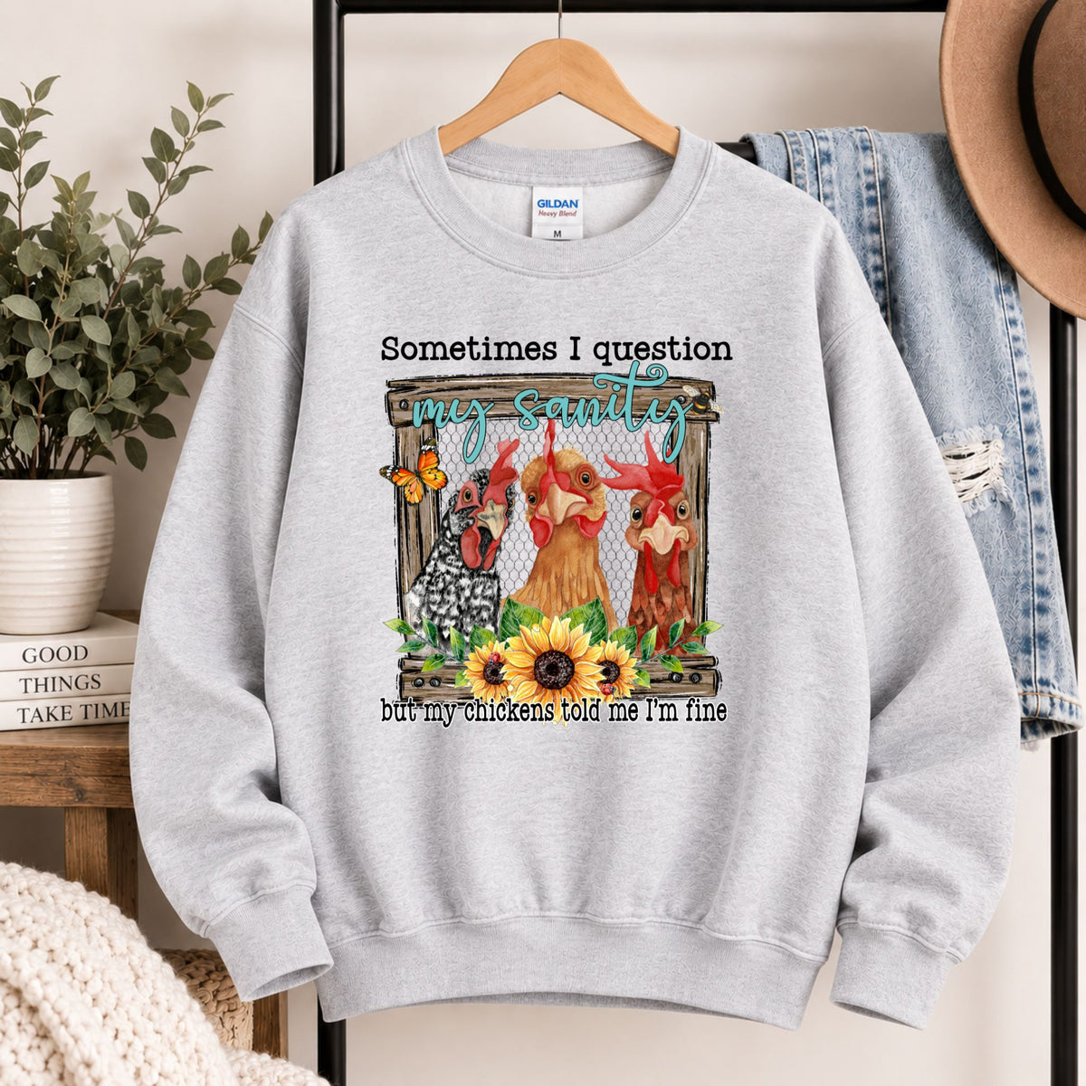 Grey crewneck sweatshirt hanging, with chicken scene graphic and quote text on front.