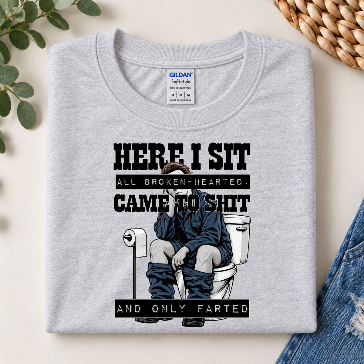 Folded light gray graphic tee with “HERE I SIT” text and toilet gag illustration.