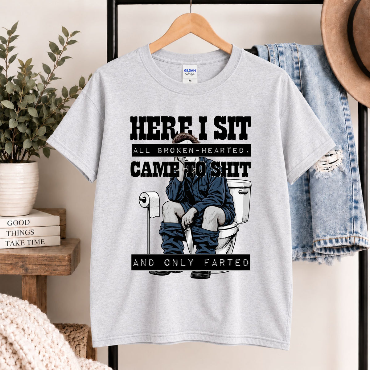 Light gray t-shirt on hanger featuring “HERE I SIT” toilet gag illustration on front.