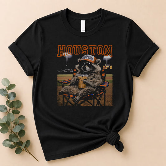 The Vintage Houston Raccoon Baseball Tee by Becca's Banging Designs LLC features a black oversized graphic shirt with a raccoon in sunglasses and an Astros cap, lounging with a drink and bold orange "HOUSTON"—perfect for game day. Beige background shown.