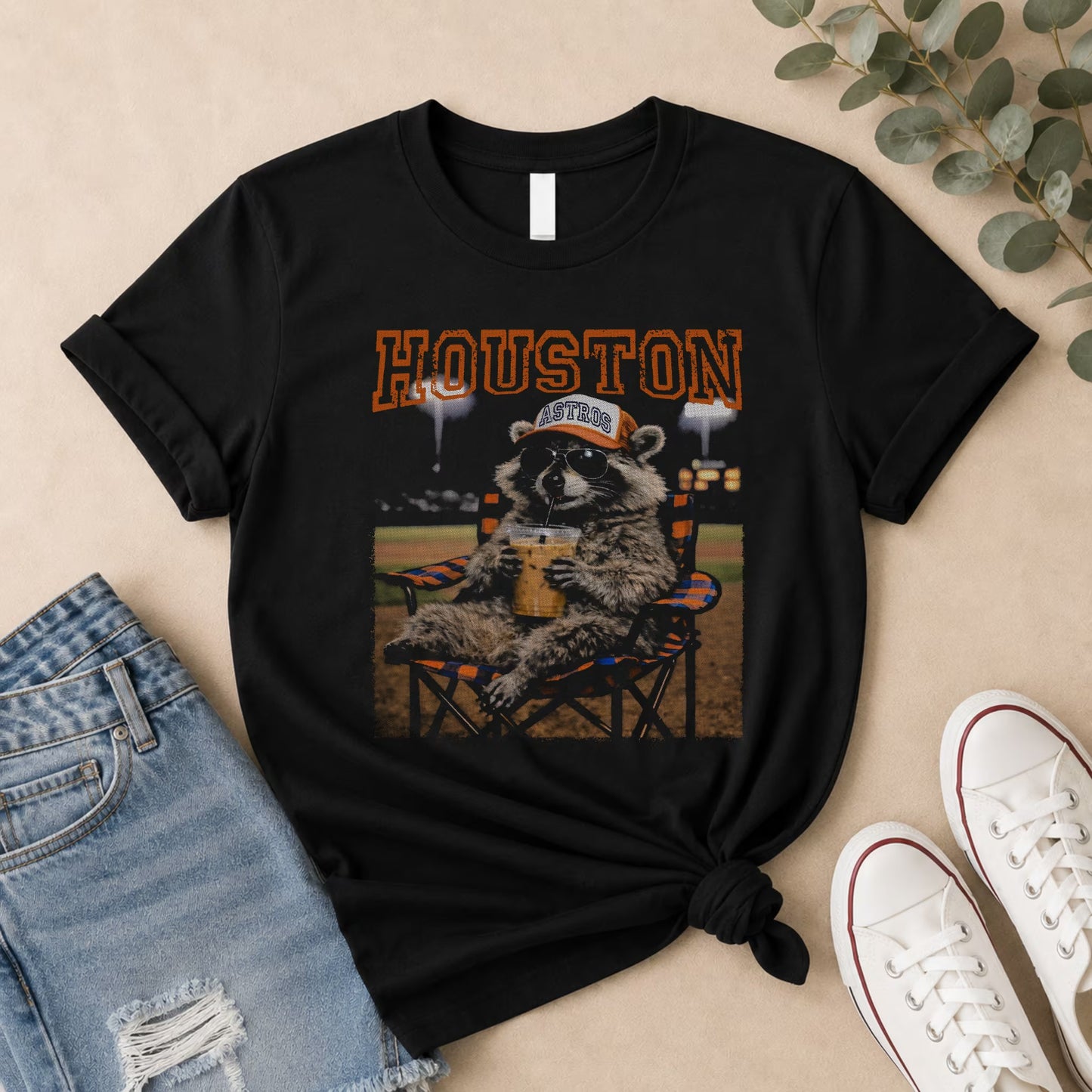 The Vintage Houston Raccoon Baseball Tee by Becca's Banging Designs LLC features a black oversized fit, HOUSTON in orange, and a raccoon in an Astros cap—ideal for game day looks with denim shorts and white sneakers.