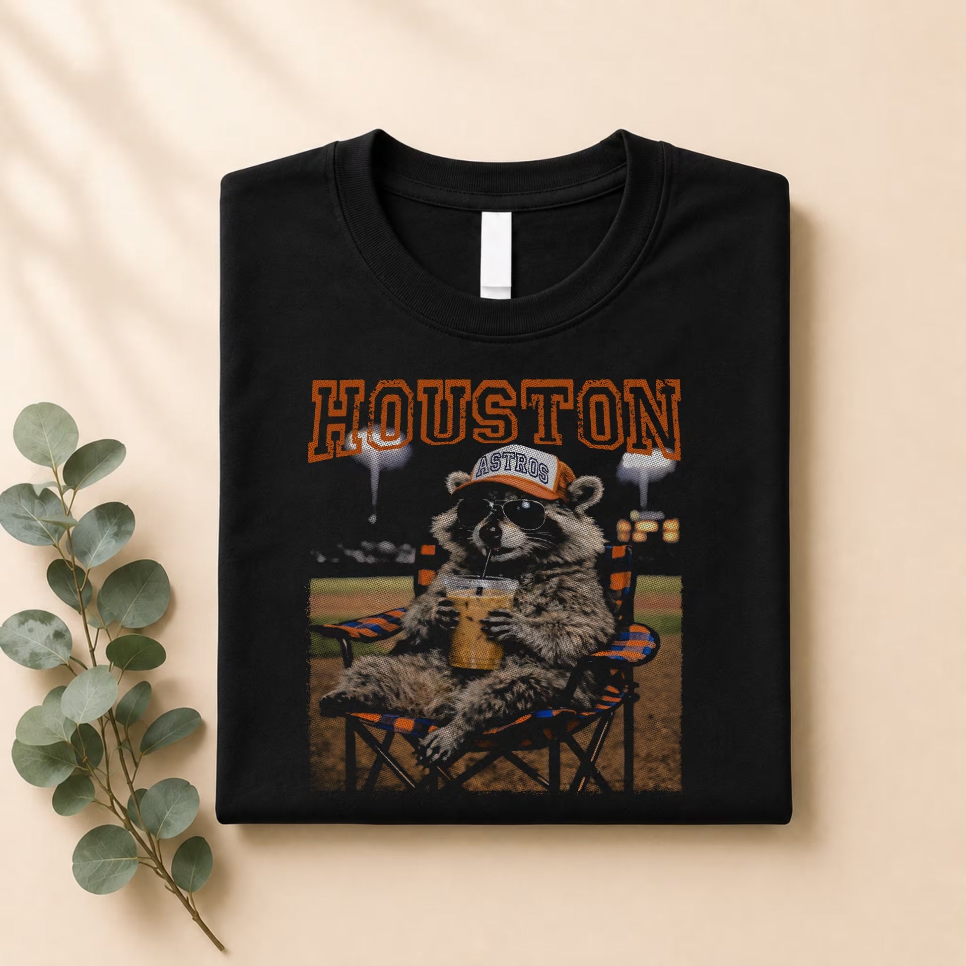 The Vintage Houston Raccoon Baseball Tee by Becca's Banging Designs LLC is a black oversized graphic shirt with a raccoon in sunglasses and an Astros cap, lounging with a drink under "Houston"—perfect for game day. Shown folded with a sprig of leaves.