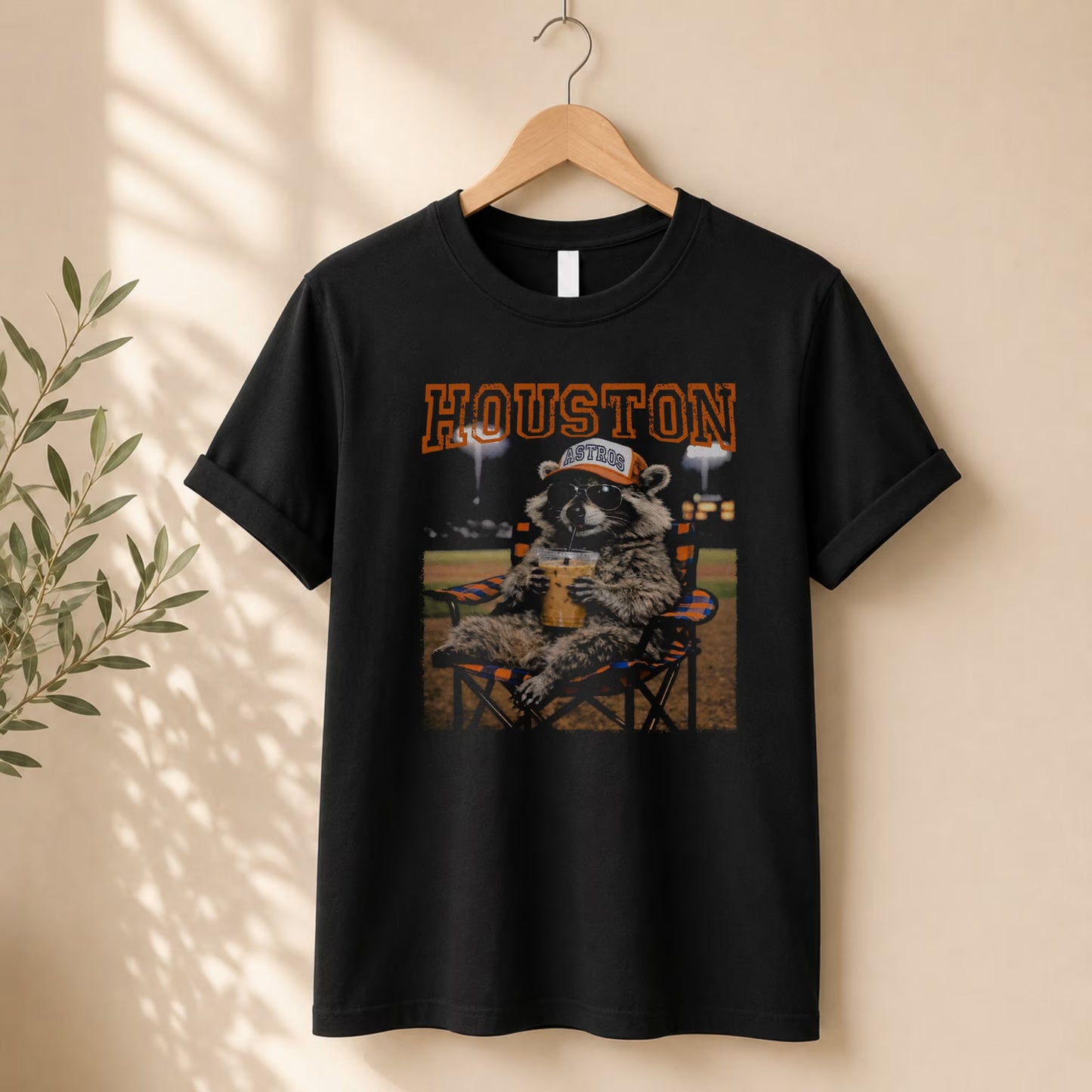 A black oversized tee from Becca's Banging Designs LLC hangs on a rack, showing a raccoon in a cap holding a drink at a baseball field with bold orange "HOUSTON”—the Vintage Houston Raccoon Baseball Tee, ideal for game day fans.