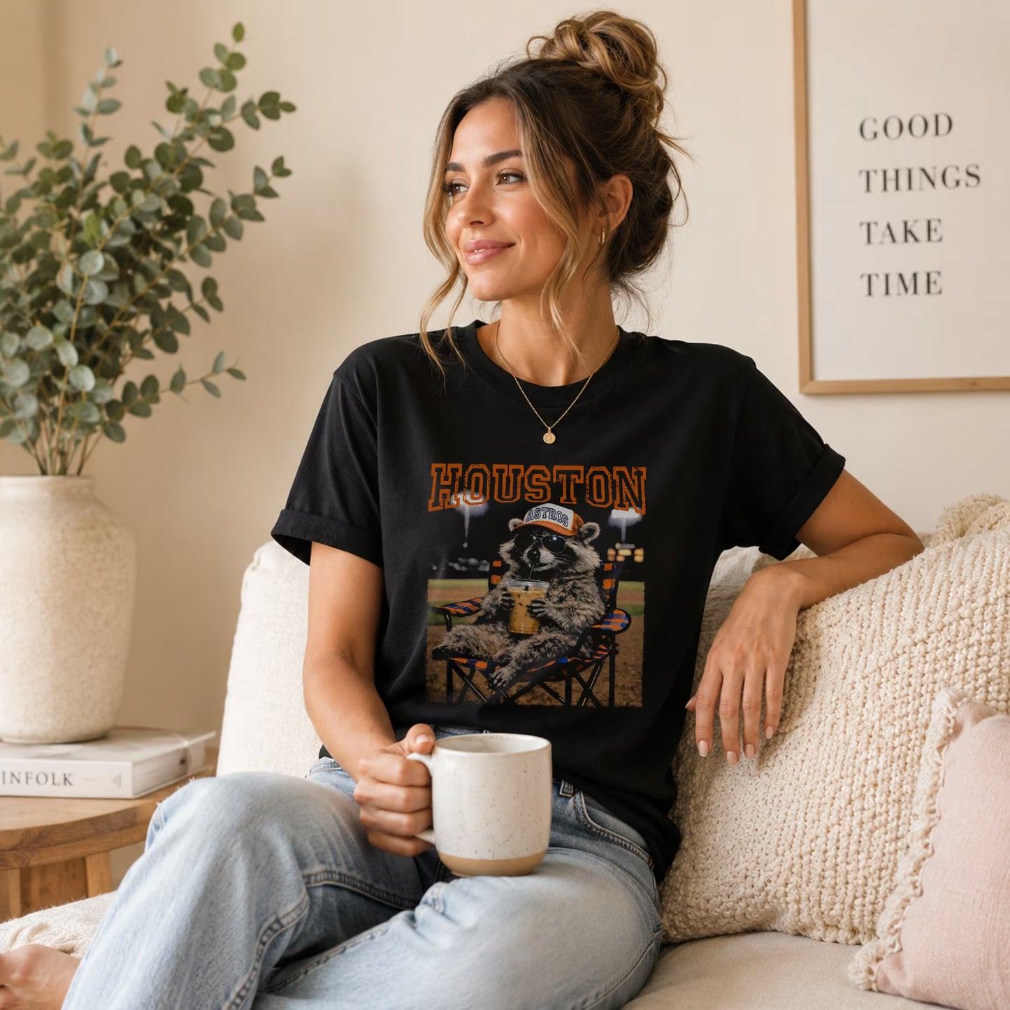 A woman with brown hair in a loose bun sits on a beige couch, smiling while holding a mug. She wears the Vintage Houston Raccoon Baseball Tee by Becca's Banging Designs LLC—an ideal relaxed game day graphic shirt. A leafy plant and framed quote are behind her.