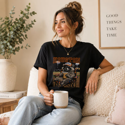 A woman with brown hair in a loose bun sits on a beige couch, smiling while holding a mug. She wears the Vintage Houston Raccoon Baseball Tee by Becca's Banging Designs LLC—an ideal relaxed game day graphic shirt. A leafy plant and framed quote are behind her.