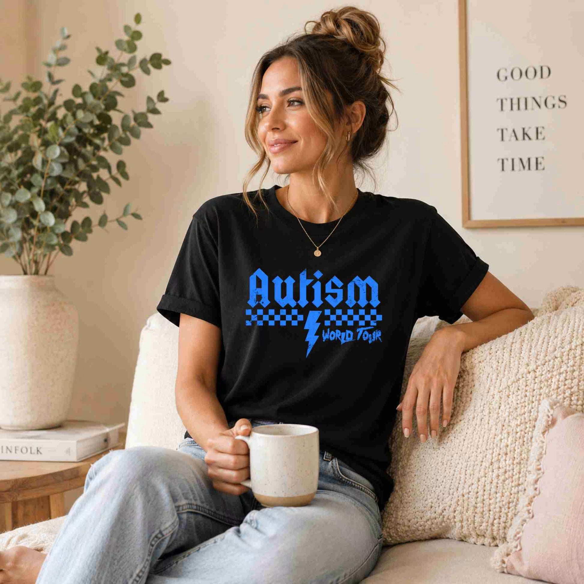 Woman wearing a black t-shirt with 'Autism World Tour' design, sitting on a couch holding a mug.