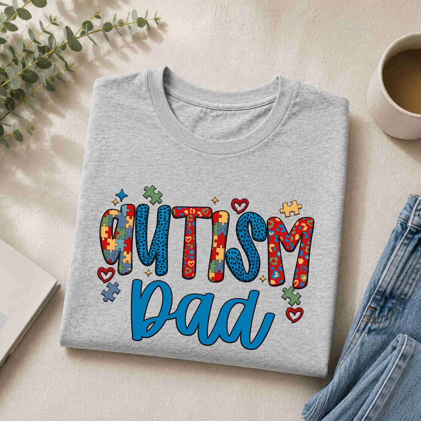 Folded gray t-shirt with 'Autism Dad' text on a light surface with a cup and denim jeans.