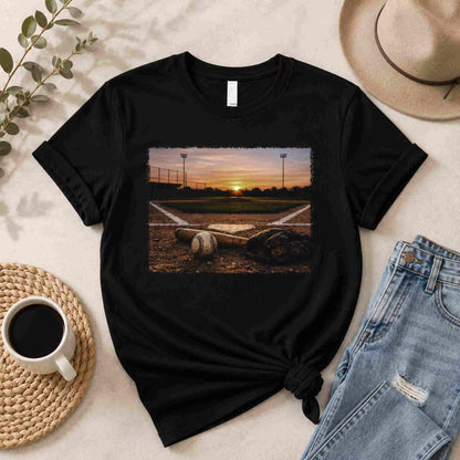 Black t-shirt with a baseball field and sunset graphic, placed on a surface with a cup, jeans, and hat.