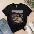 The Softball Mom Iced Coffee Bleacher Queen Graphic Tee by Becca's Banging Designs LLC features a smiling woman outdoors with a drink—a fun, trendy choice for softball moms and fans of women’s graphic t-shirts.