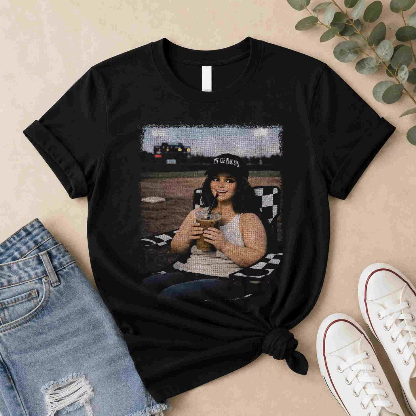 The Softball Mom Iced Coffee Bleacher Queen Graphic Tee by Becca's Banging Designs LLC features a smiling woman outdoors with a drink—a fun, trendy choice for softball moms and fans of women’s graphic t-shirts.