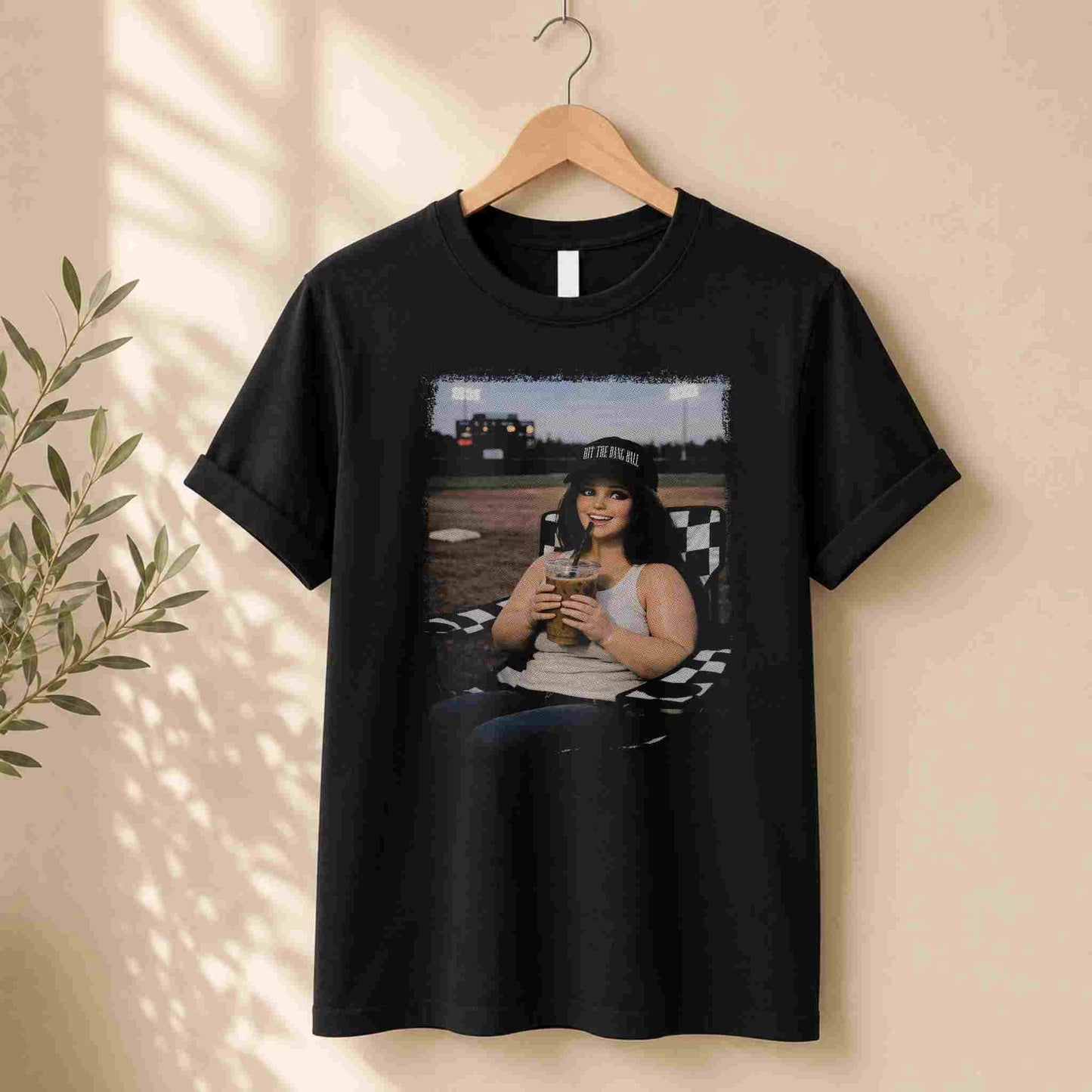 A black, affordable Becca's Banging Designs LLC tee hangs on a wooden hanger, featuring the "Softball Mom Iced Coffee Bleacher Queen" graphic—a funny and trendy shirt for softball mamas to wear on any occasion.