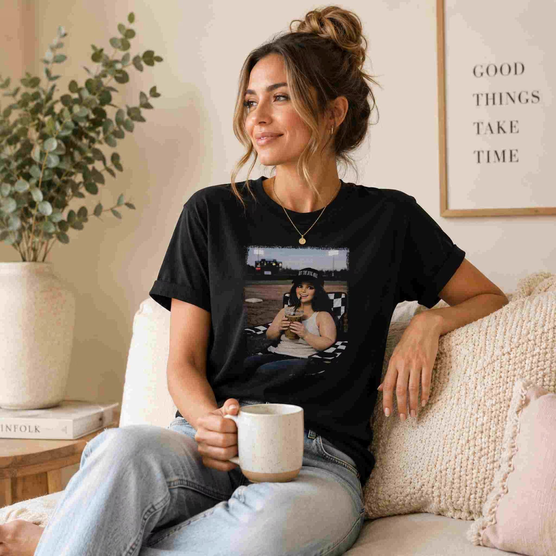 A woman with light brown hair in a loose bun sits on a beige sofa, smiling with a mug. She wears the Becca's Banging Designs LLC Softball Mom Iced Coffee Bleacher Queen Graphic Tee, light jeans, and gold jewelry. A sign behind her reads GOOD THINGS TAKE TIME.