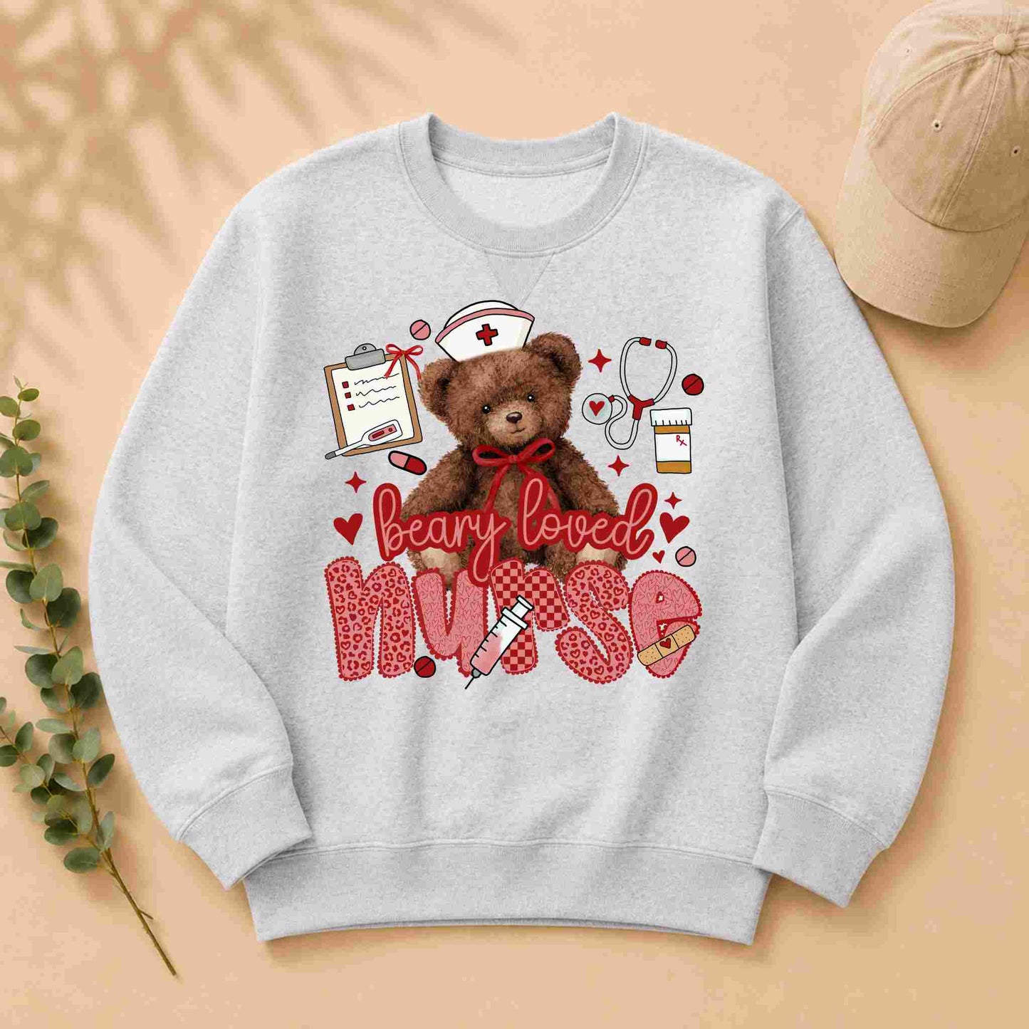 The Beary Loved Nurse Sweatshirt Comfort Style and Care by Becca's Banging Designs features a cute teddy bear in a nurse hat with medical items and hearts, plus playful "beary loved nurse" text—an ideal gift for nurses.