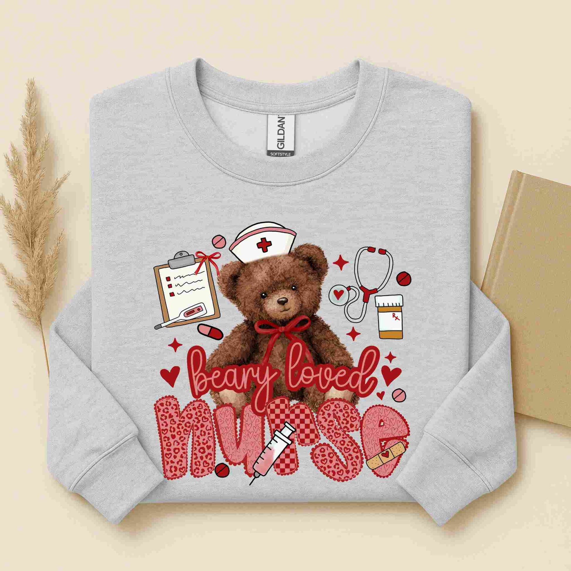 The Beary Loved Nurse Sweatshirt by Becca's Banging Designs features a cute teddy bear in a nurse’s hat with medical items and hearts, plus "nurse" in pink leopard print—a cozy, thoughtful gift for nurses.