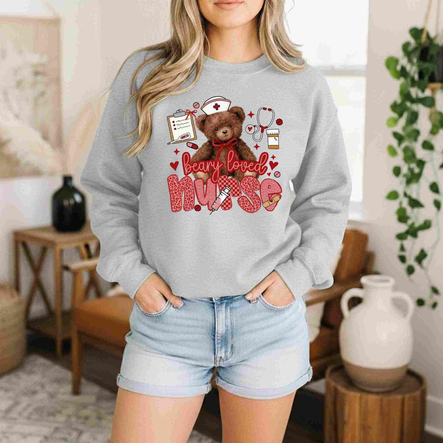 Wearing the Beary Loved Nurse Sweatshirt Comfort Style and Care by Becca's Banging Designs, a woman stands indoors with hands in her jean shorts pockets. This cozy gray crewneck features a teddy bear, nurse icons, and "beary loved nurse" in red—a perfect nurse gift.
