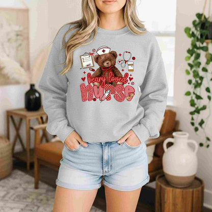 Wearing the Beary Loved Nurse Sweatshirt Comfort Style and Care by Becca's Banging Designs, a woman stands indoors with hands in her jean shorts pockets. This cozy gray crewneck features a teddy bear, nurse icons, and "beary loved nurse" in red—a perfect nurse gift.