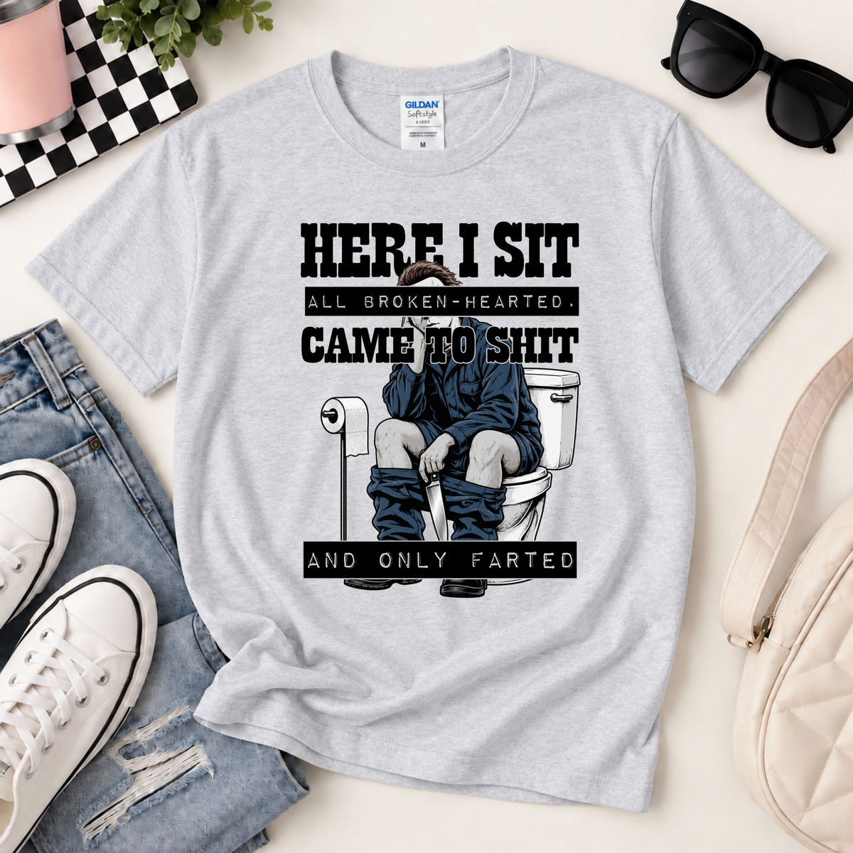 Light gray graphic tee laid flat with toilet gag print reading “HERE I SIT”.