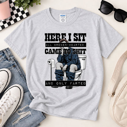 Light gray graphic tee laid flat with toilet gag print reading “HERE I SIT”.