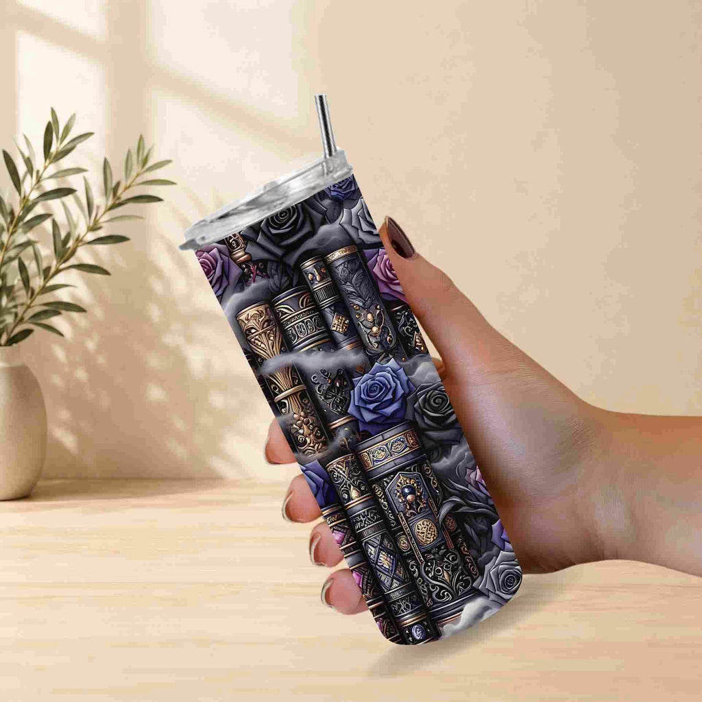 A hand holds the Dark Book Lover 20oz Skinny Tumbler from Becca's Banging Designs LLC, featuring vintage books and purple-blue roses. A potted plant sits behind on a light surface—an ideal gift for book lovers.