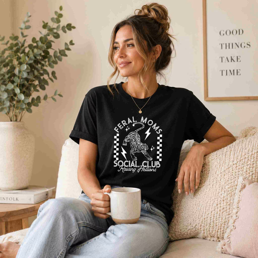 A woman relaxes on a couch with a mug, wearing the Feral Moms Social Club – Raising Hellions Graphic Tee by Becca's Banging Designs LLC. A framed sign behind her reads, “Good things take time.”.