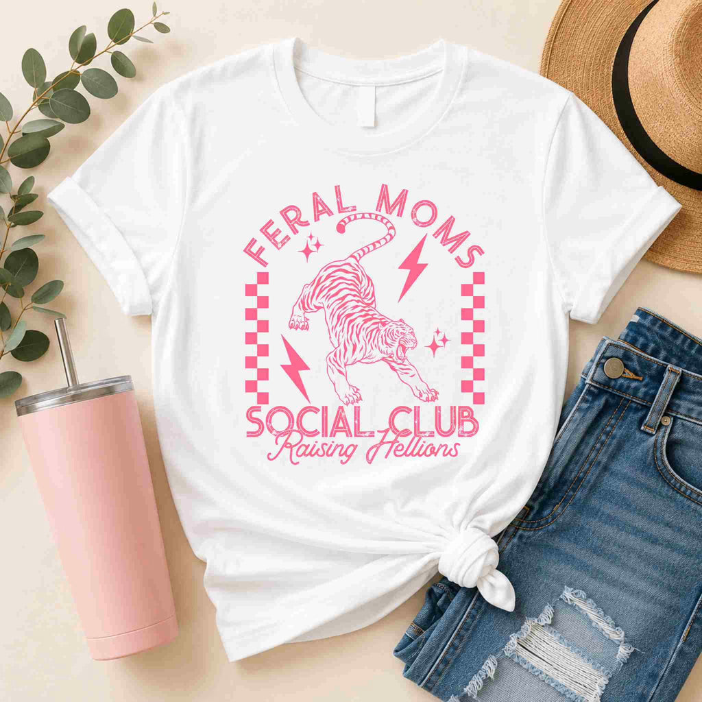 The Feral Moms Social Club Shirt – Raising Hellions Graphic Tee by Becca's Banging Designs LLC features pink text and a tiger graphic on white. Pair it with denim shorts, a tan hat, and a pink tumbler for a playful, casual look.