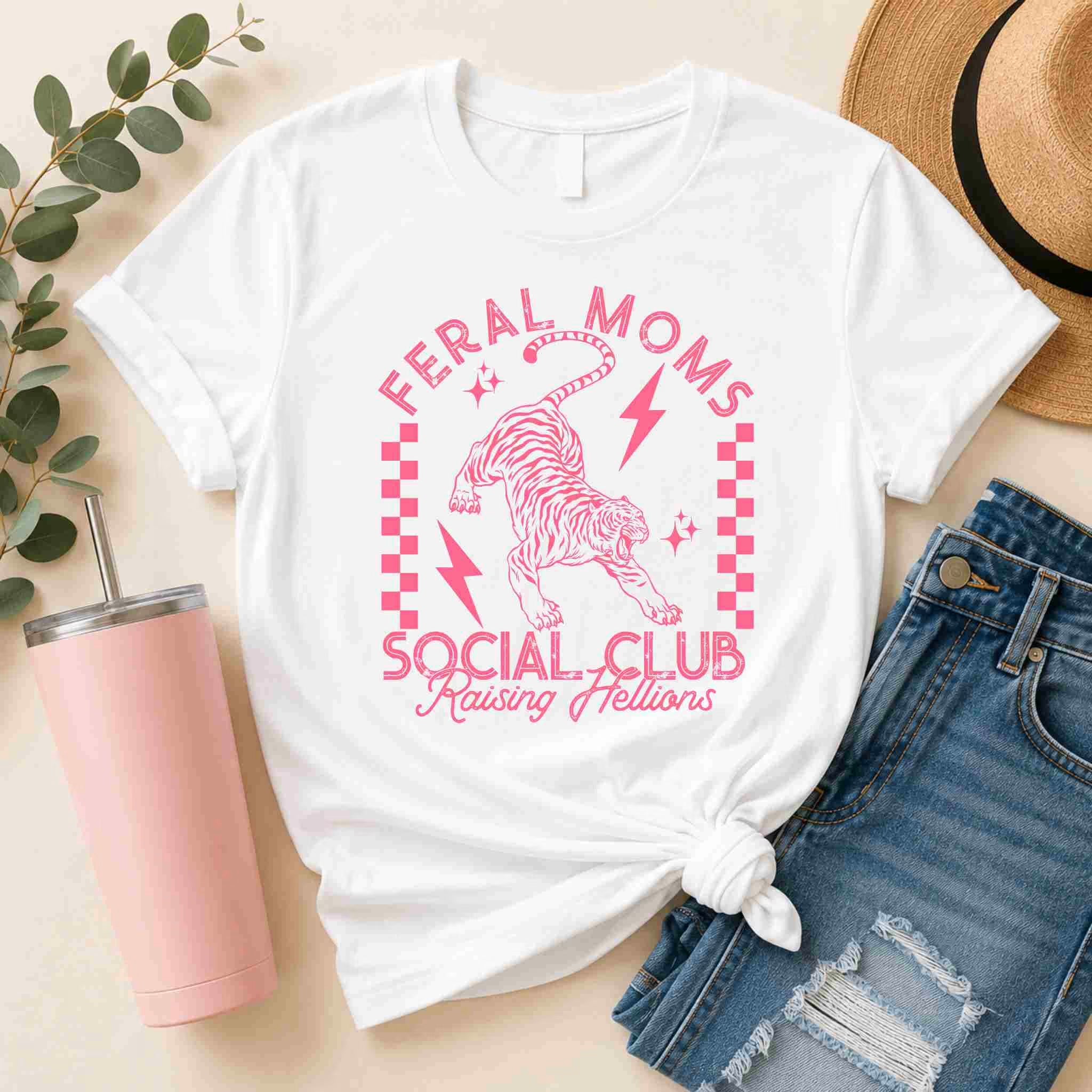 The Feral Moms Social Club Shirt – Raising Hellions Graphic Tee by Becca's Banging Designs LLC features pink text and a tiger graphic on white. Pair it with denim shorts, a tan hat, and a pink tumbler for a playful, casual look.