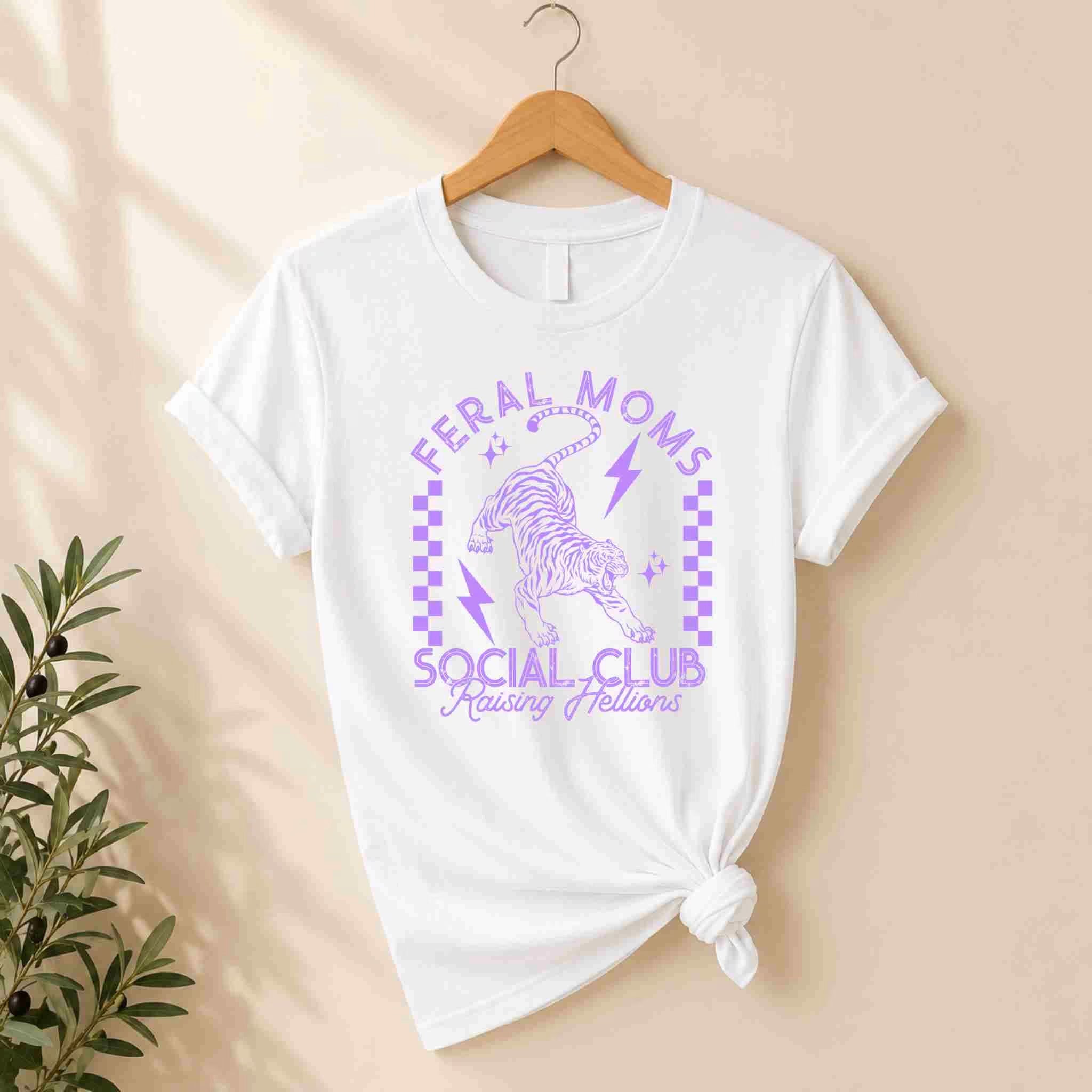 A white Feral Moms Social Club Shirt – Raising Hellions Graphic Tee by Becca's Banging Designs LLC hangs on display, featuring a purple tiger, lightning bolts, stars, and a checkerboard design. A plant appears on the left for added charm.