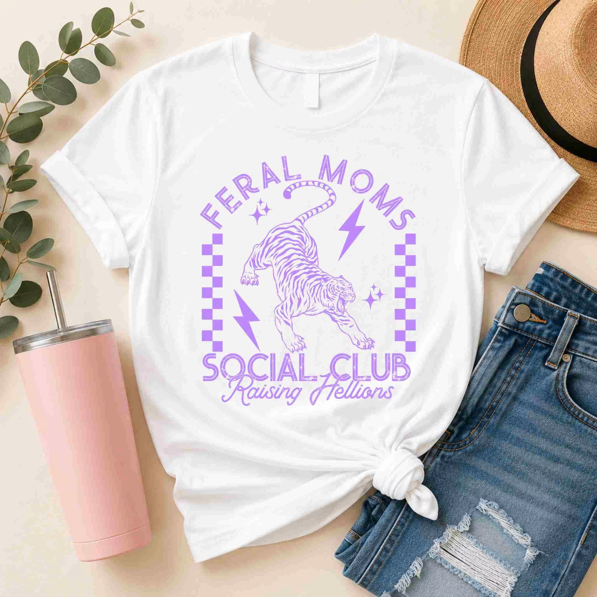 The Feral Moms Social Club Shirt – Raising Hellions Graphic Tee by Becca's Banging Designs LLC features bold purple text, a tiger, lightning bolts, and checkerboard accents—styled with denim shorts and a straw hat for summer flair.