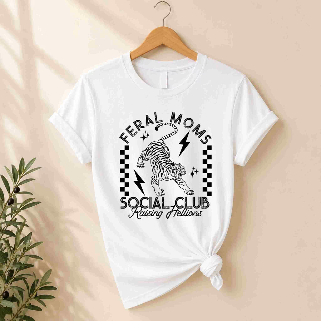 The Feral Moms Social Club Shirt – Raising Hellions Graphic Tee by Becca's Banging Designs LLC hangs on a wooden hanger, featuring a bold tiger graphic, black lightning bolts, checkerboard patterns, and “Raising Hellions” text. A plant sits to the left for style.