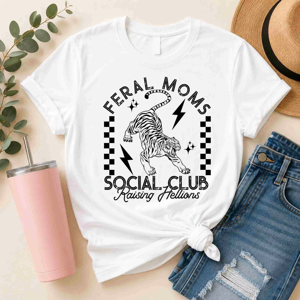 The Feral Moms Social Club Shirt – Raising Hellions Graphic Tee by Becca's Banging Designs LLC features a bold tiger graphic and text, styled with ripped shorts, a straw hat, pink tumbler, and leafy decor for an eye-catching look.