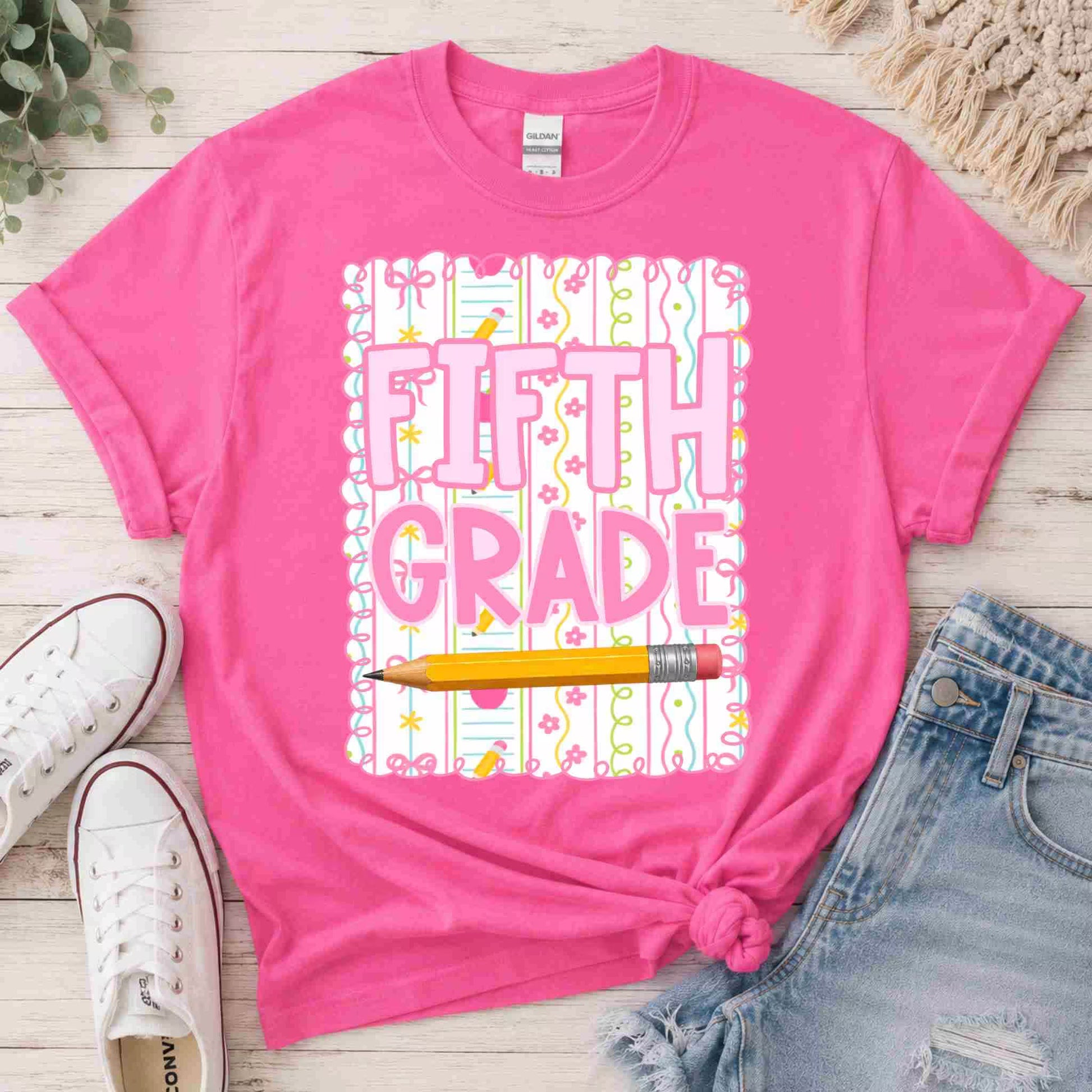 Pink t-shirt with 'Fifth Grade' design on a wooden surface with shoes and shorts.