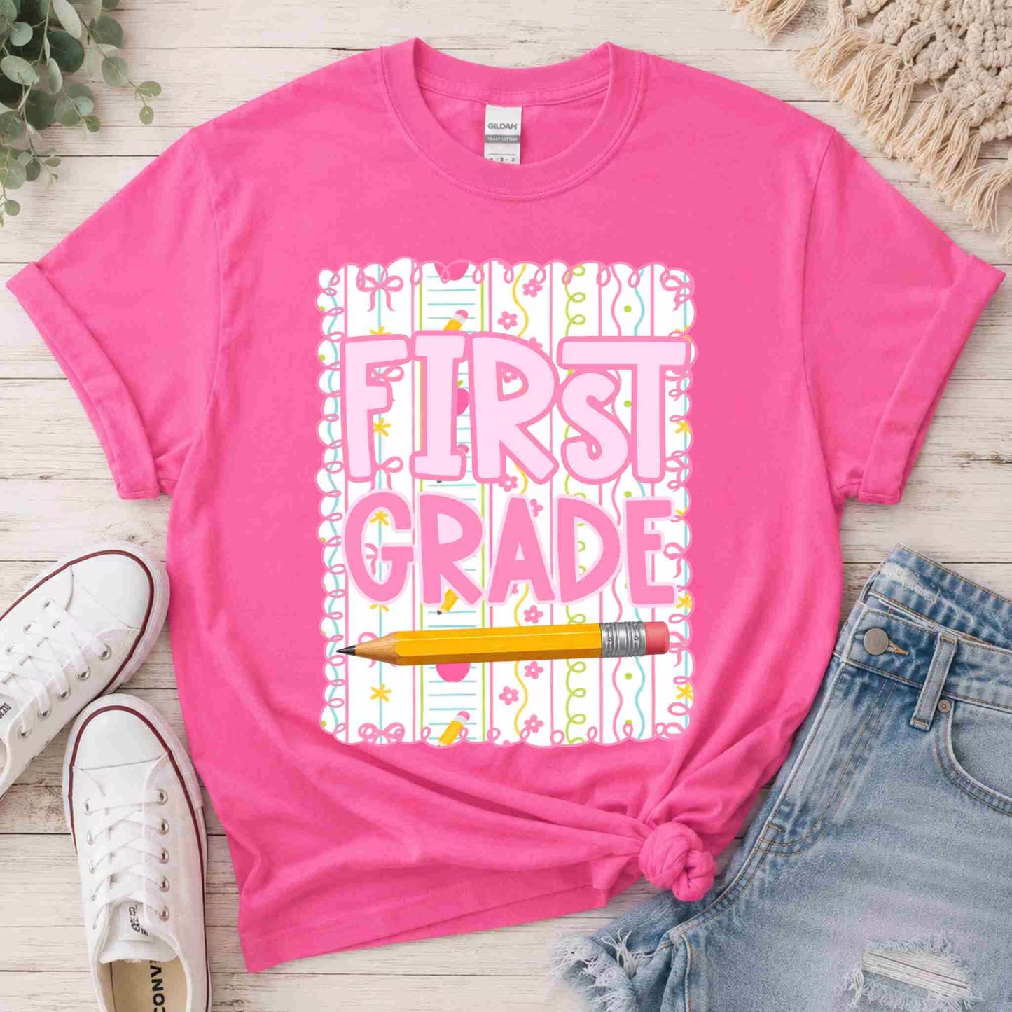 Pink t-shirt with 'First Grade' design on a wooden surface with shoes and jeans.