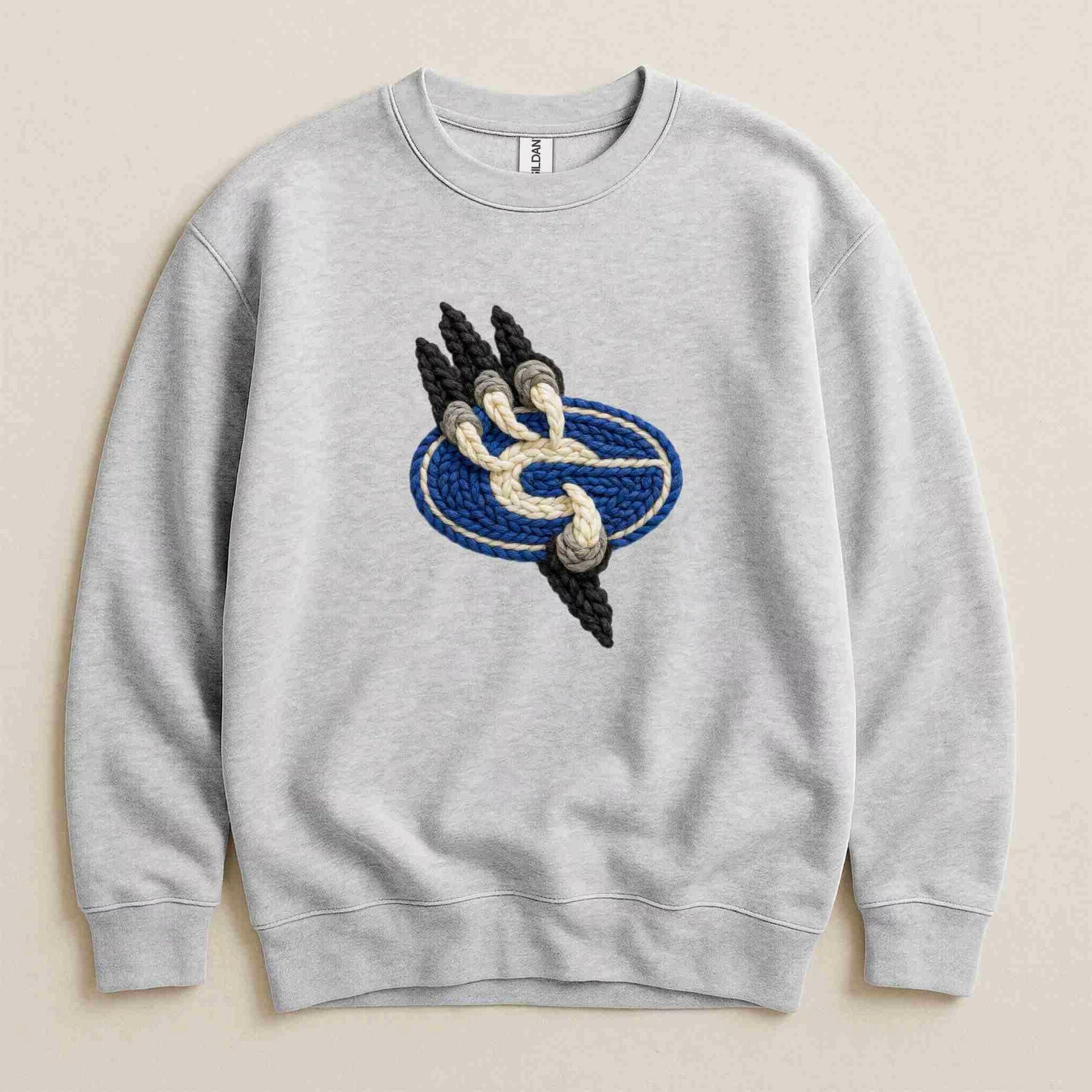 The Gateway Griffins | Crewneck by Becca's Banging Designs is a cozy, oversized light gray sweatshirt featuring a central embroidered patch with a blue oval, beige rope accent, and three black spikes on the left.