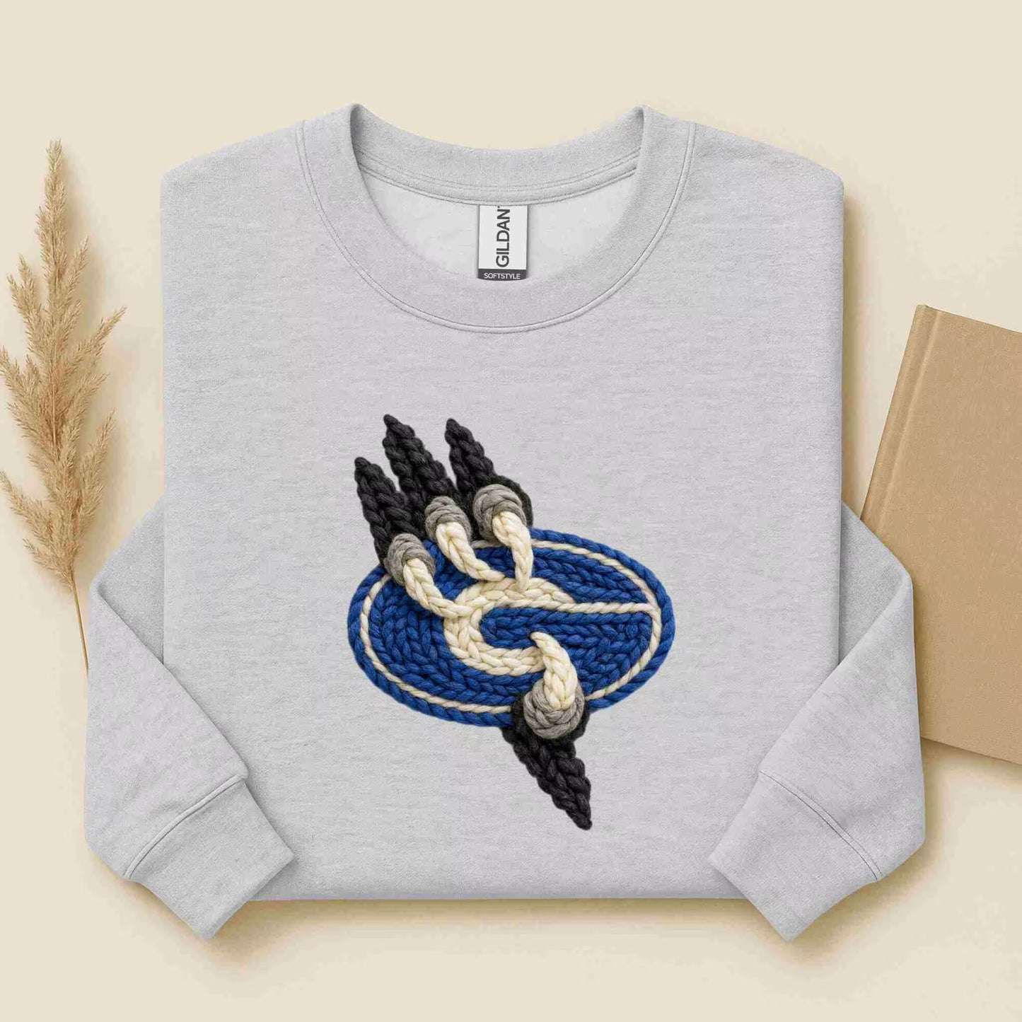 The Gateway Griffins | Crewneck by Becca's Banging Designs is a light gray, oversized sweatshirt featuring a textured blue oval and embroidered paw print in black, gray, and cream yarn. Shown on beige with a wheat stalk and brown book nearby.