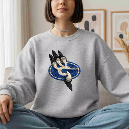 A woman sits cross-legged indoors wearing the Gateway Griffins Crewneck by Becca's Banging Designs, featuring a blue oval and knot design. With chin-length hair, a simple choker, and abstract art behind her, she exudes cozy style.