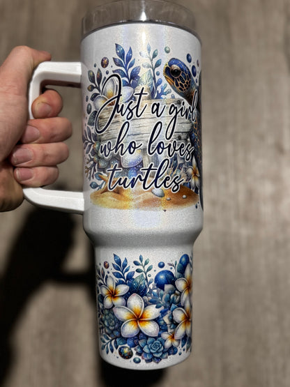 A hand holds Becca's Banging Designs LLC’s “Just a Girl Who Loves Turtles” Glitter Tumbler, a 40oz floral beach cup with handle featuring seashells, flowers, and a turtle on a white background against a wooden surface.