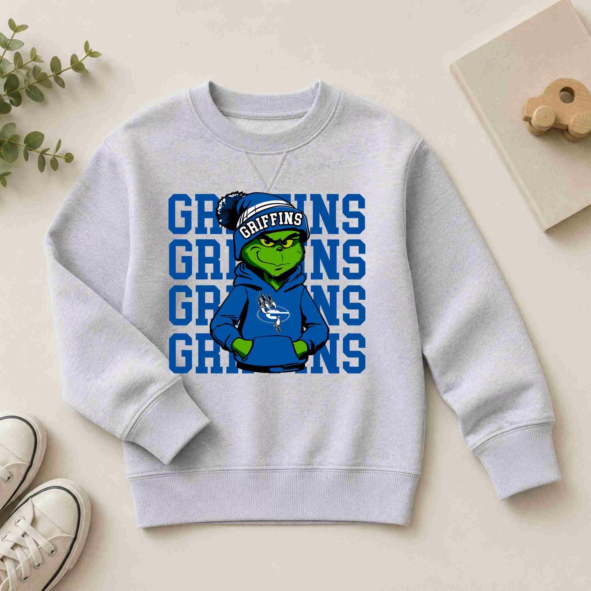 The Griffins Crewneck Sweatshirt Youth Comfort and Style by Becca's Banging Designs features a green cartoon griffin in a blue hoodie and hat with bold blue "GRIFFINS" text—perfect for school spirit. Shoes, a plant, and a wooden toy are nearby.