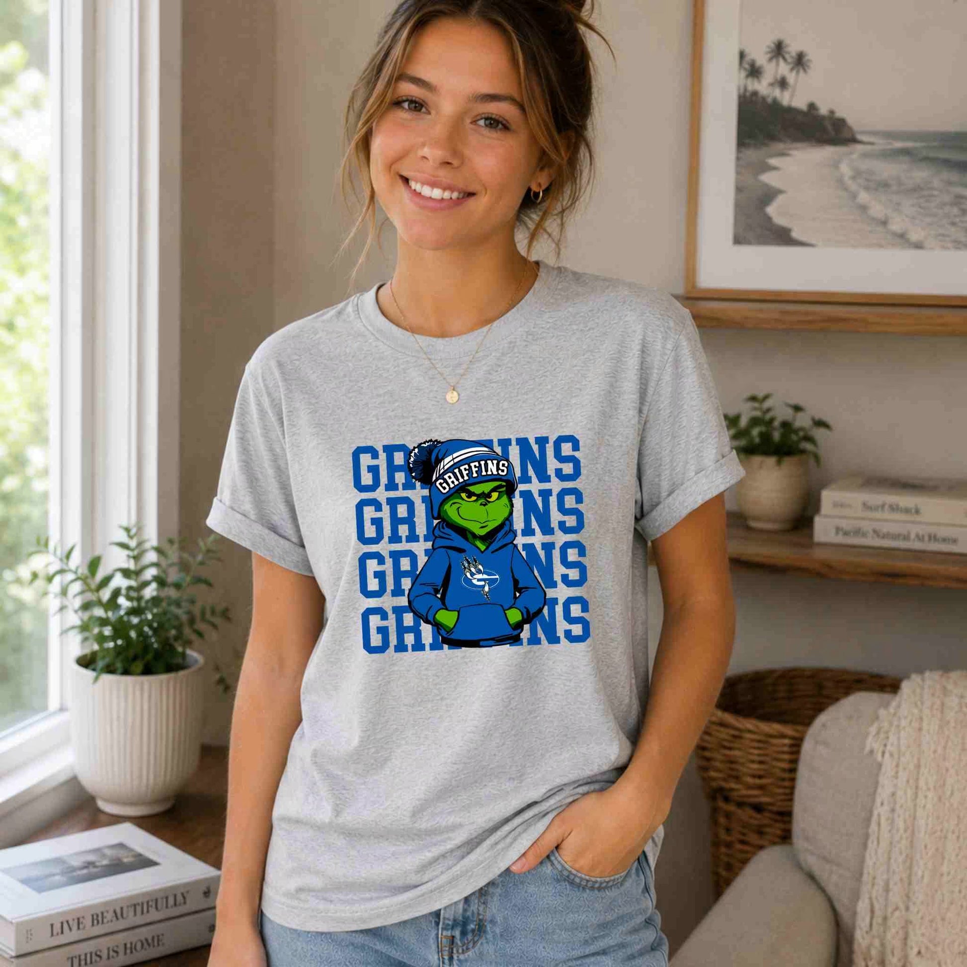 A smiling woman in a cozy room wears the Griffins Graphic Tee Youth Comfort and School Spirit by Becca's Banging Designs, featuring a cartoon green griffin and bold logo, with plants, books, and beach art showing her school pride.