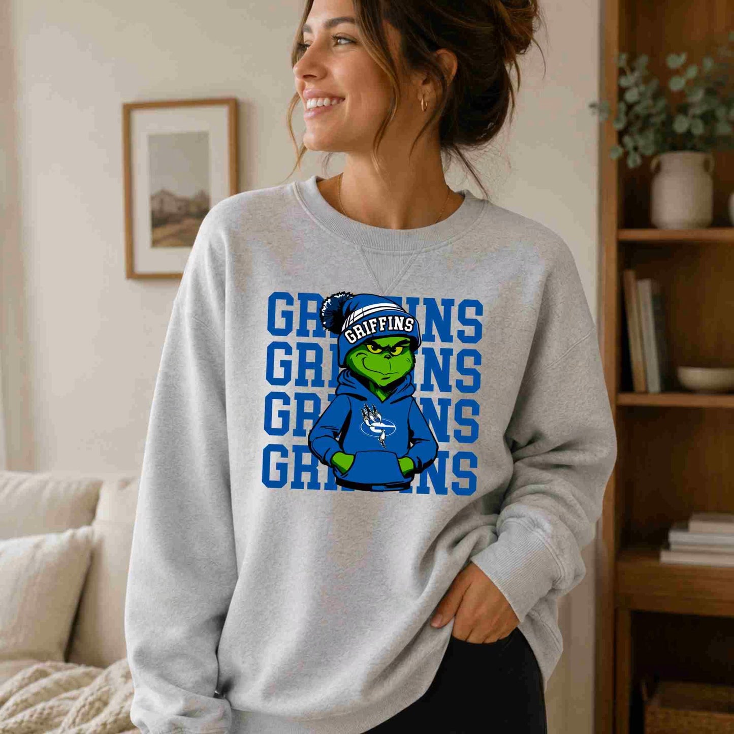 A woman smiles indoors, wearing Becca's Banging Designs' Griffins Crewneck Sweatshirt Youth Comfort and Style, which features a cartoon green griffin in a blue hoodie and headband before repeated blue "GRIFFINS" text.