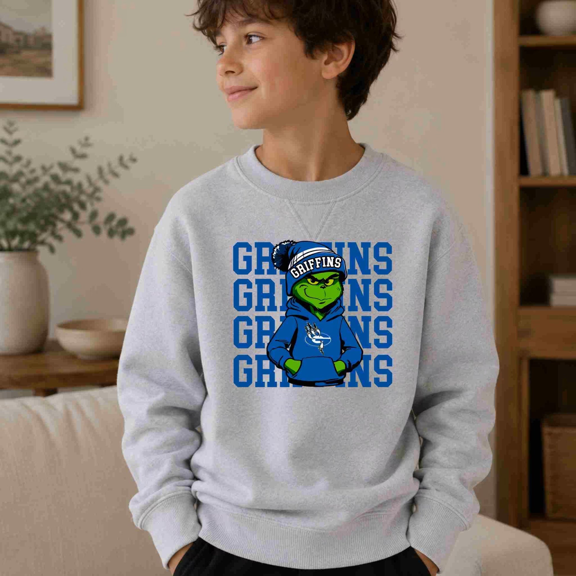 A smiling boy wears the Griffins Crewneck Sweatshirt Youth Comfort and Style by Becca's Banging Designs, featuring a cartoon frog in a blue hoodie and GRIFFINS cap with repeated blue GRIFFINS text, showing his school spirit indoors.