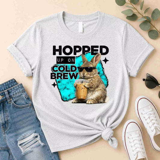 The Hopped Up On Cold Brew Bunny Shirt by Becca's Banging Designs LLC is a light gray tee with a rabbit in sunglasses holding cold brew and the phrase “Hopped Up On Cold Brew”—perfect for coffee lovers.