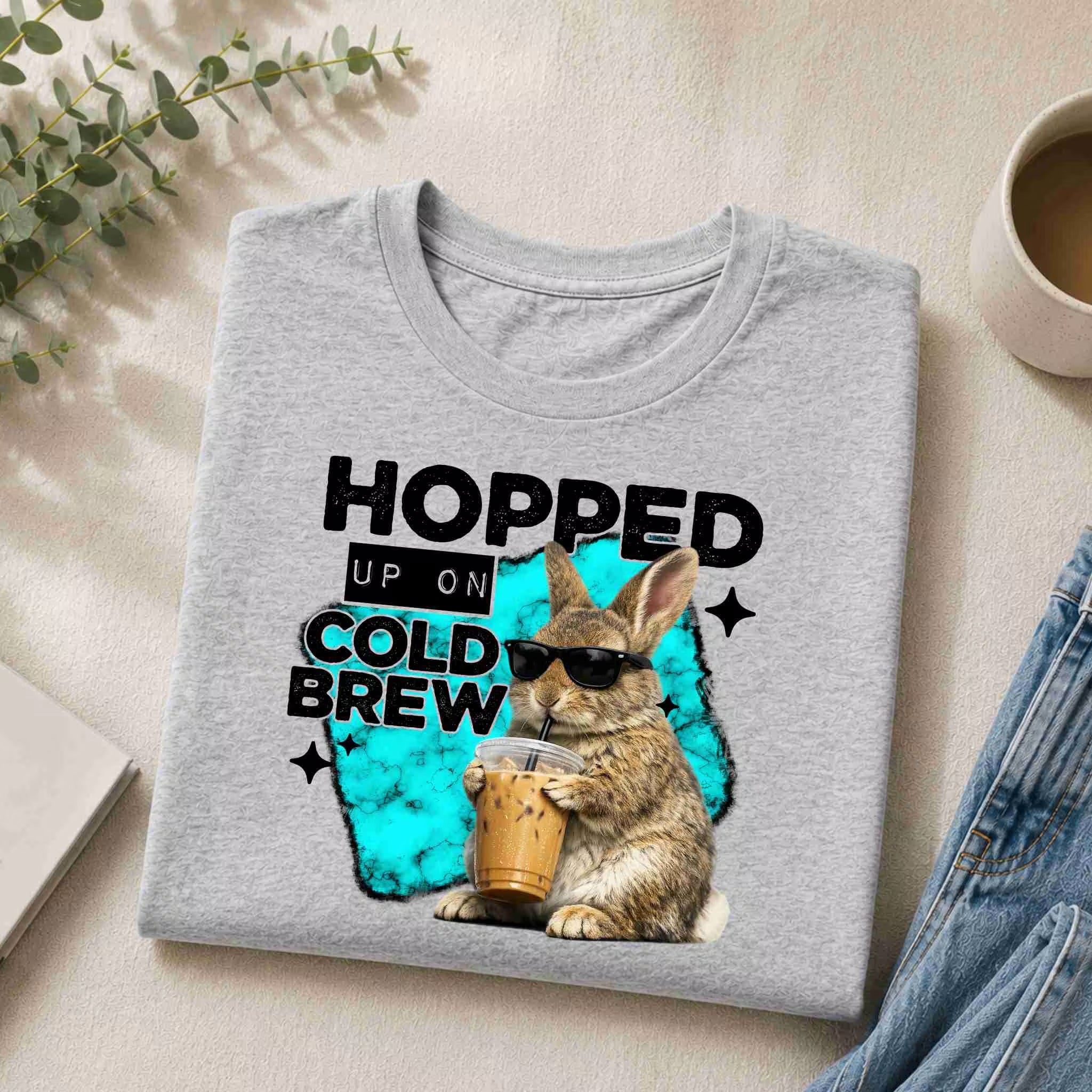 The Hopped Up On Cold Brew Bunny Shirt by Becca's Banging Designs LLC is a gray tee featuring a cold brew bunny in sunglasses holding an iced coffee, with "Hopped Up On Cold Brew" text in black and blue—perfect for coffee lovers.