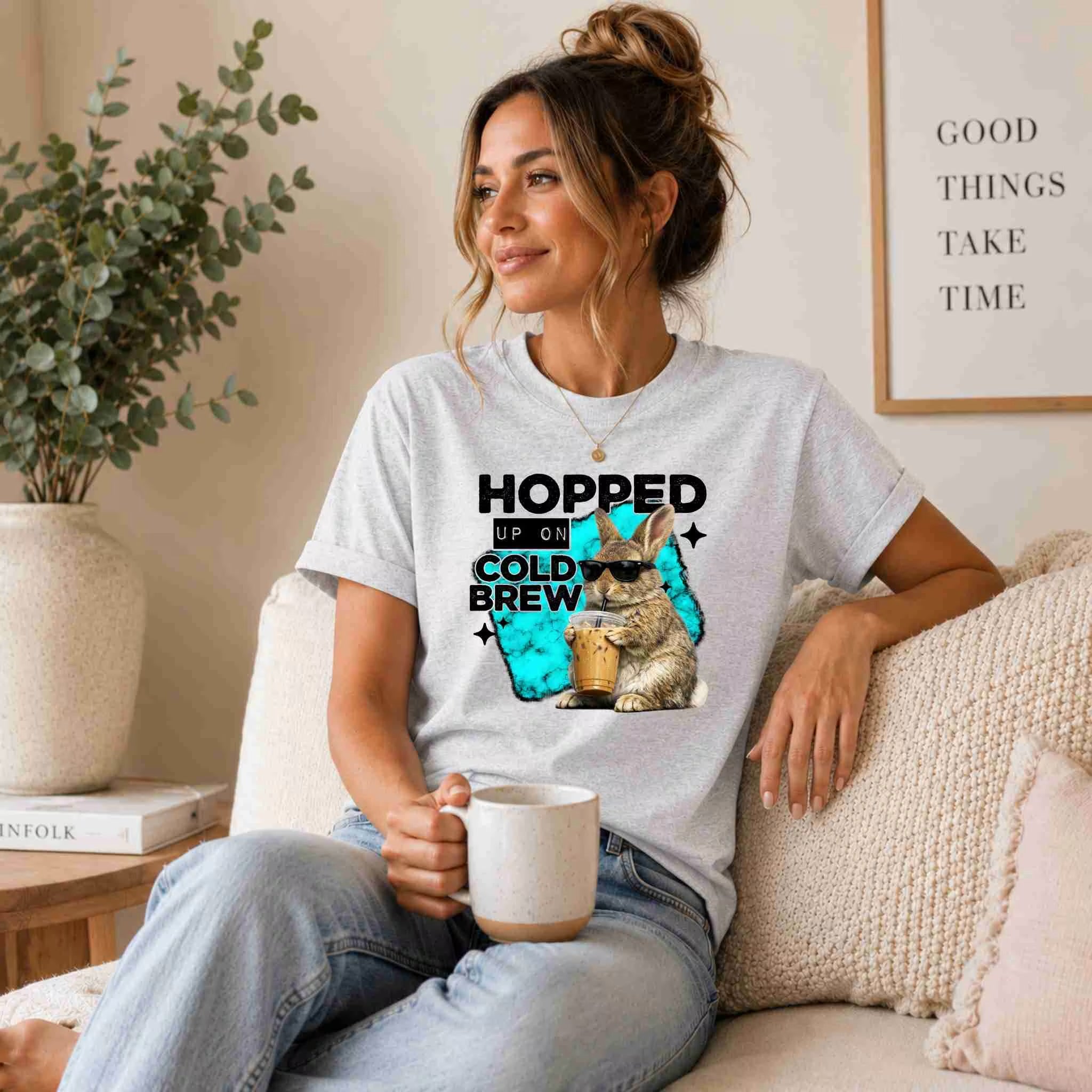 A woman sits on a beige couch holding a coffee mug, wearing the Hopped Up On Cold Brew Bunny Shirt by Becca's Banging Designs LLC; behind her, a wall sign reads "Good things take time.