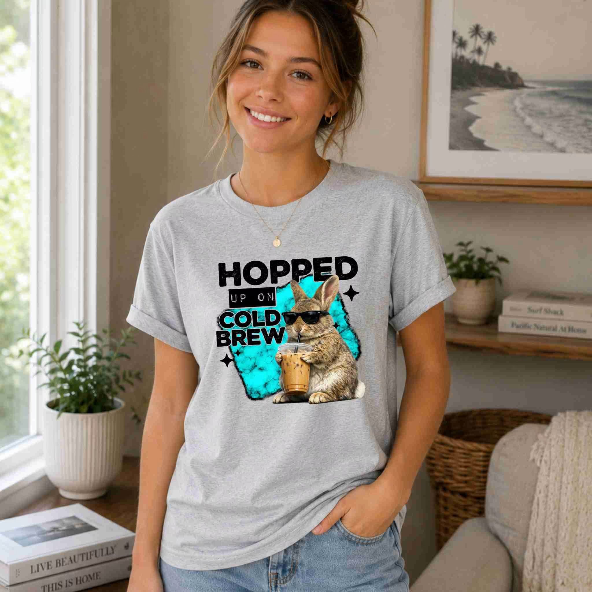 A woman smiles indoors wearing the “Hopped Up On Cold Brew Bunny Shirt” by Becca's Banging Designs LLC, a quirky coffee graphic tee. Houseplants and beachy decor add charm—ideal for fans of unique tees.
