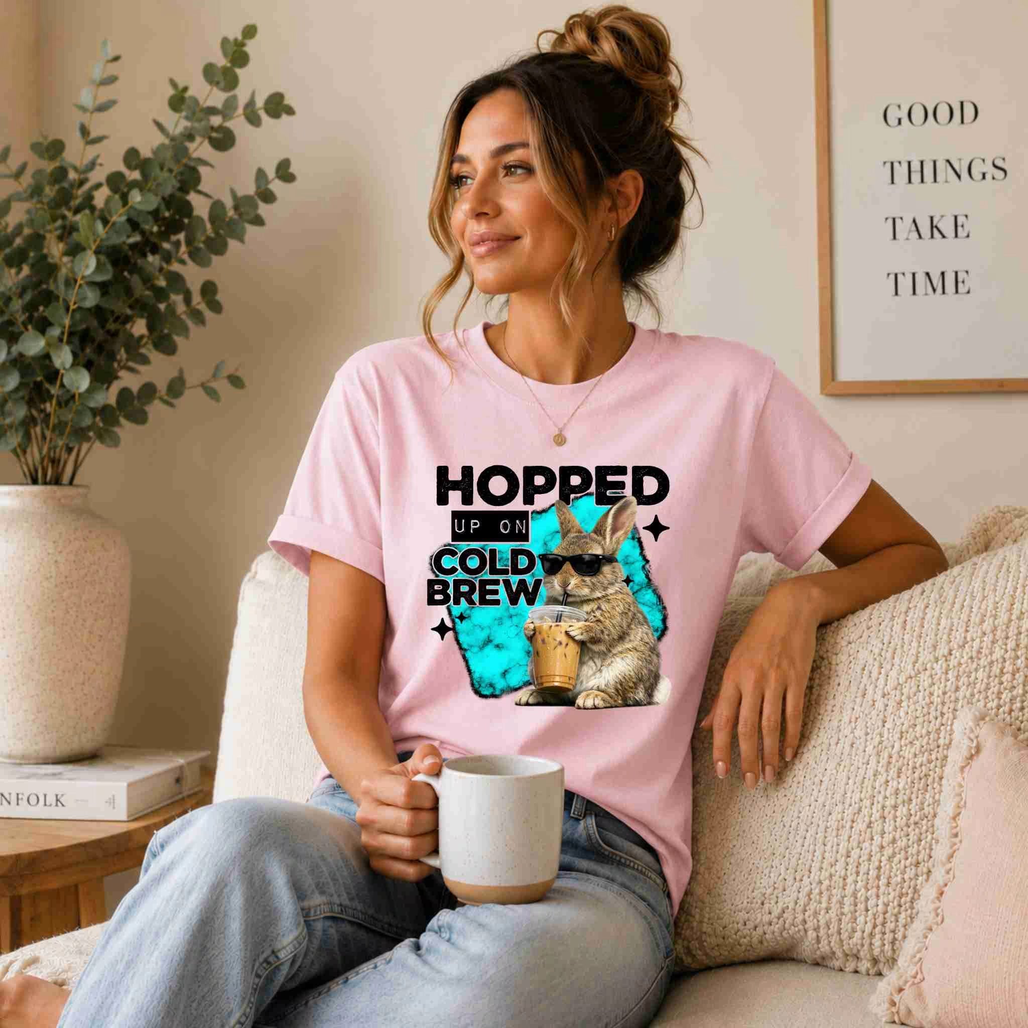 A woman smiles on a beige sofa, holding a mug and wearing the "Hopped Up On Cold Brew Bunny Shirt" by Becca's Banging Designs LLC—a funny coffee graphic tee with a cold brew bunny. A sign behind her reads "Good Things Take Time.