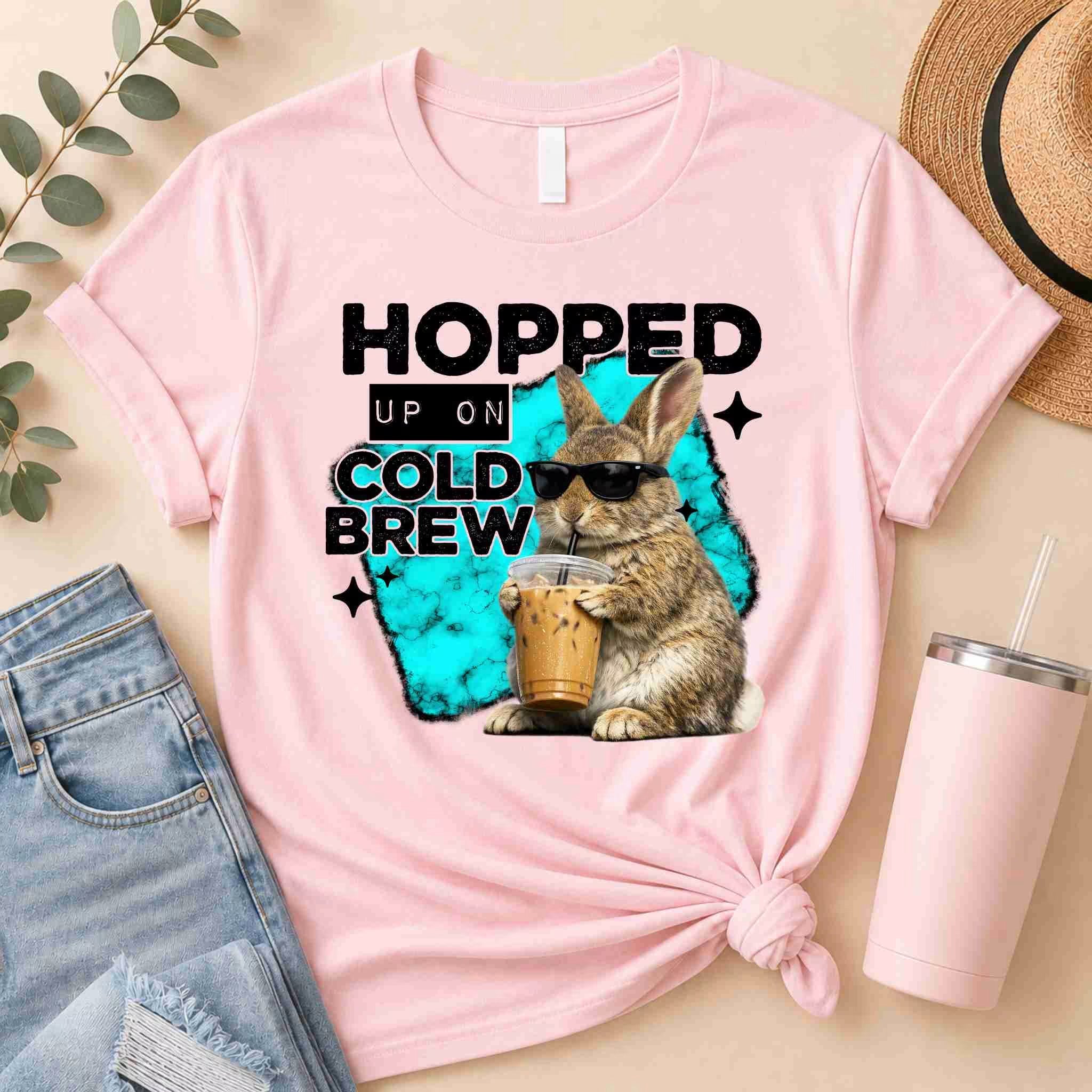 The Hopped Up On Cold Brew Bunny Shirt by Becca's Banging Designs LLC is a fun pink tee with a sunglass-wearing bunny. Perfect for coffee lovers and cute graphic T-shirt fans, it pairs well with jeans and adds playful style to any outfit.