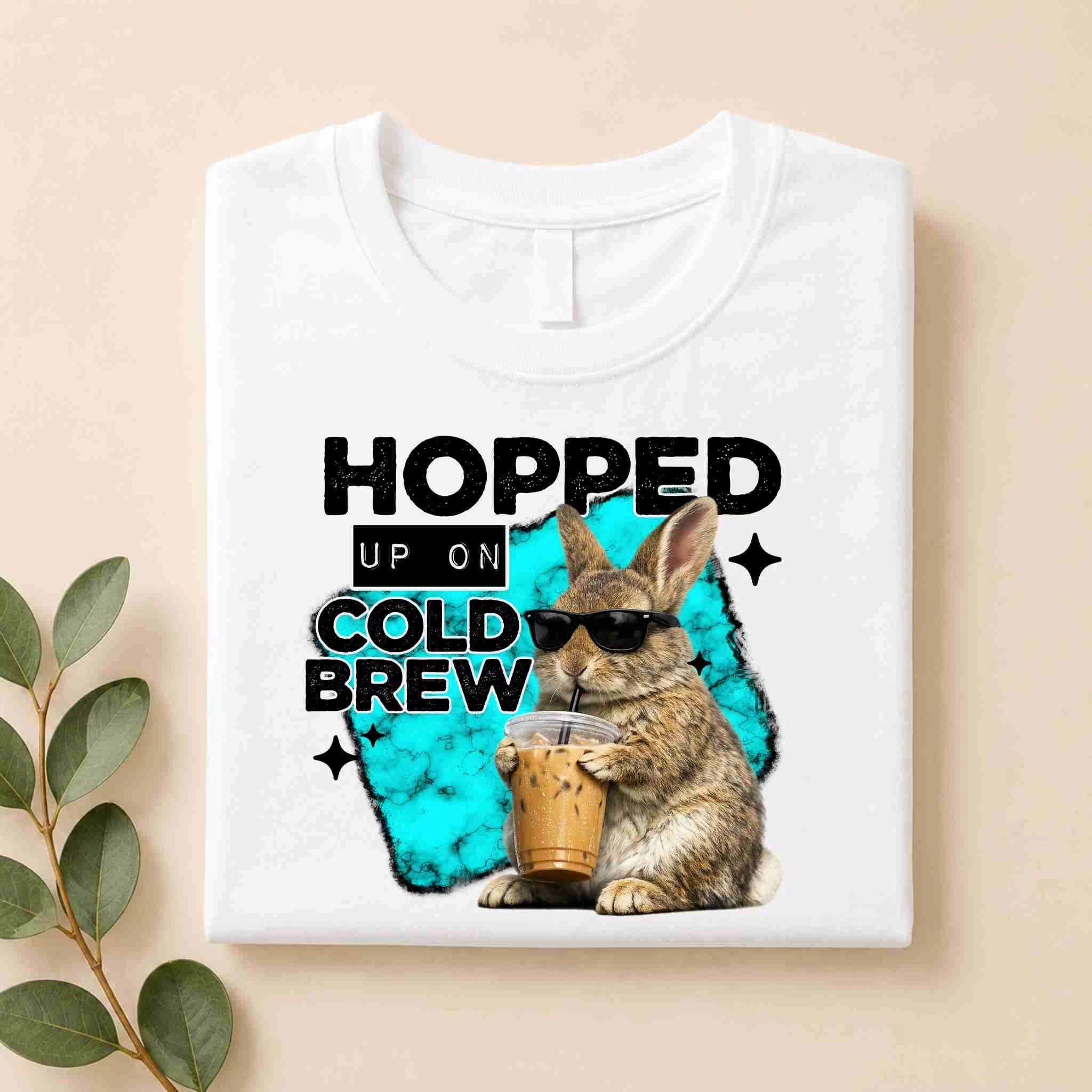 The Hopped Up On Cold Brew Bunny Shirt by Becca's Banging Designs LLC features a bunny in sunglasses holding iced coffee with "Hopped up on cold brew" text and a blue splash background, perfect for coffee lovers. A leafy branch sits beside the folded white tee.